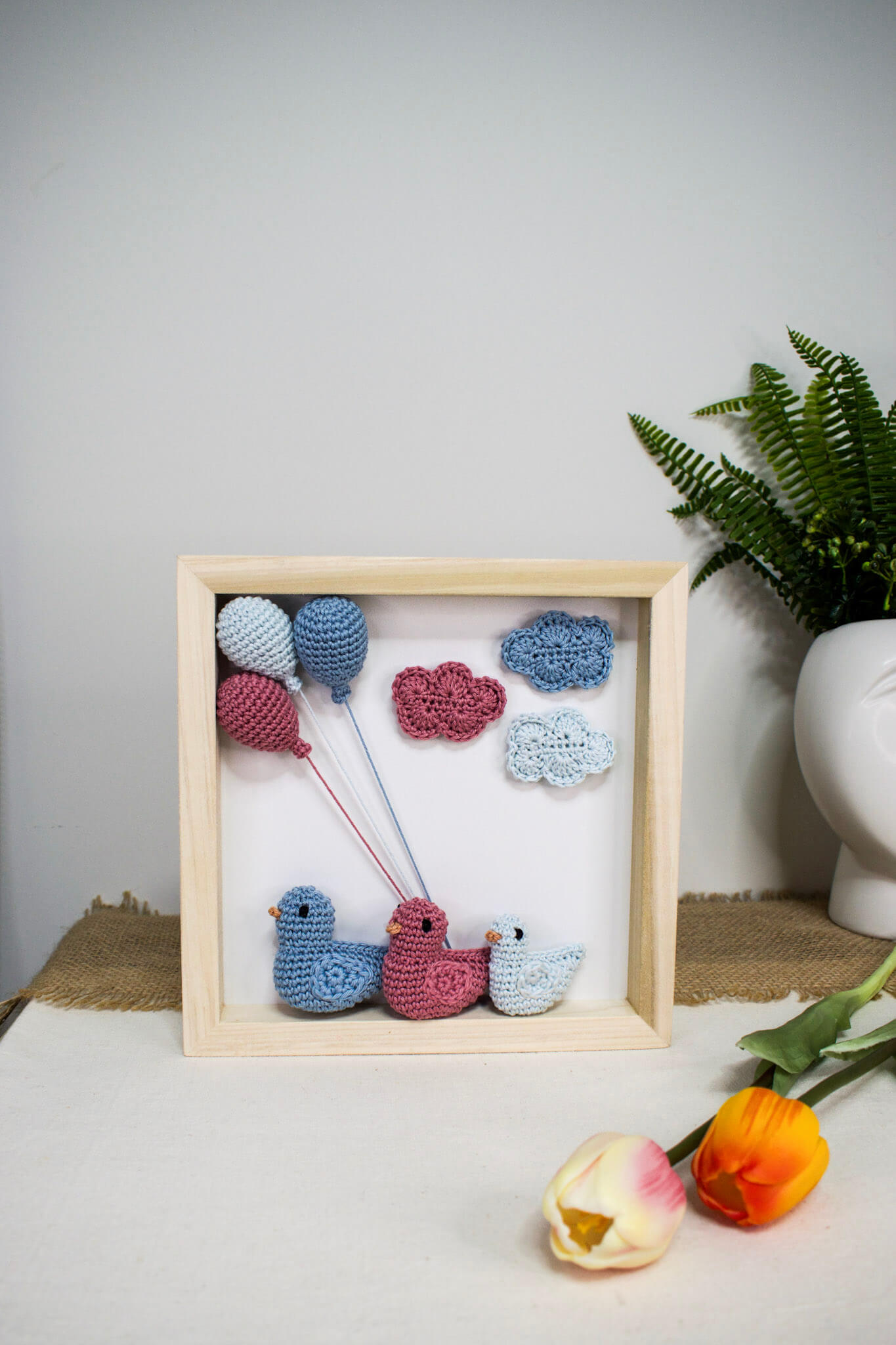 Χειροποίητο ξύλινο κάδρο με πλεκτά amigurumi παπάκια που αναπαριστούν μια οικογένεια με αγοράκι, διακοσμημένο με γαλάζια και ροζ πλεκτά μπαλόνια και σύννεφα.